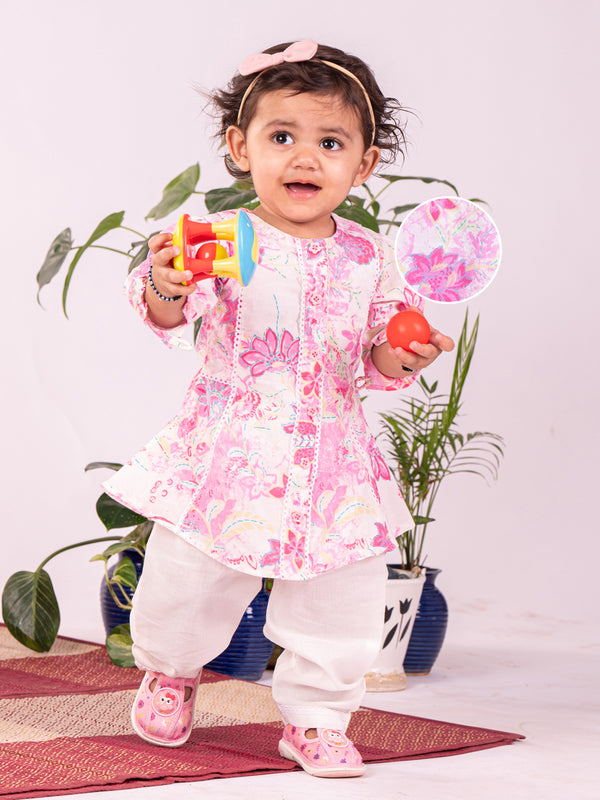 A toddler wearing a pink knee-length kurta set with a round neck and button opening, holding a toy, accompanied by a potted plant in the background.