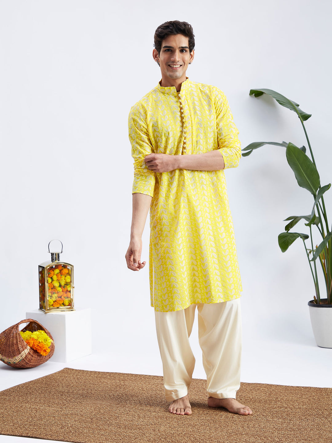 A man standing and posing in a mustard yellow chikankari embroidered Kurta with Cream Patiala set. The Kurta has long sleeves and a mandarin collar, and the patiala has an elasticated waistband.
