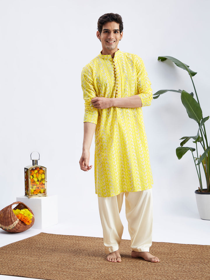 A man standing and posing in a mustard yellow chikankari embroidered Kurta with Cream Patiala set. The Kurta has long sleeves and a mandarin collar, and the patiala has an elasticated waistband.