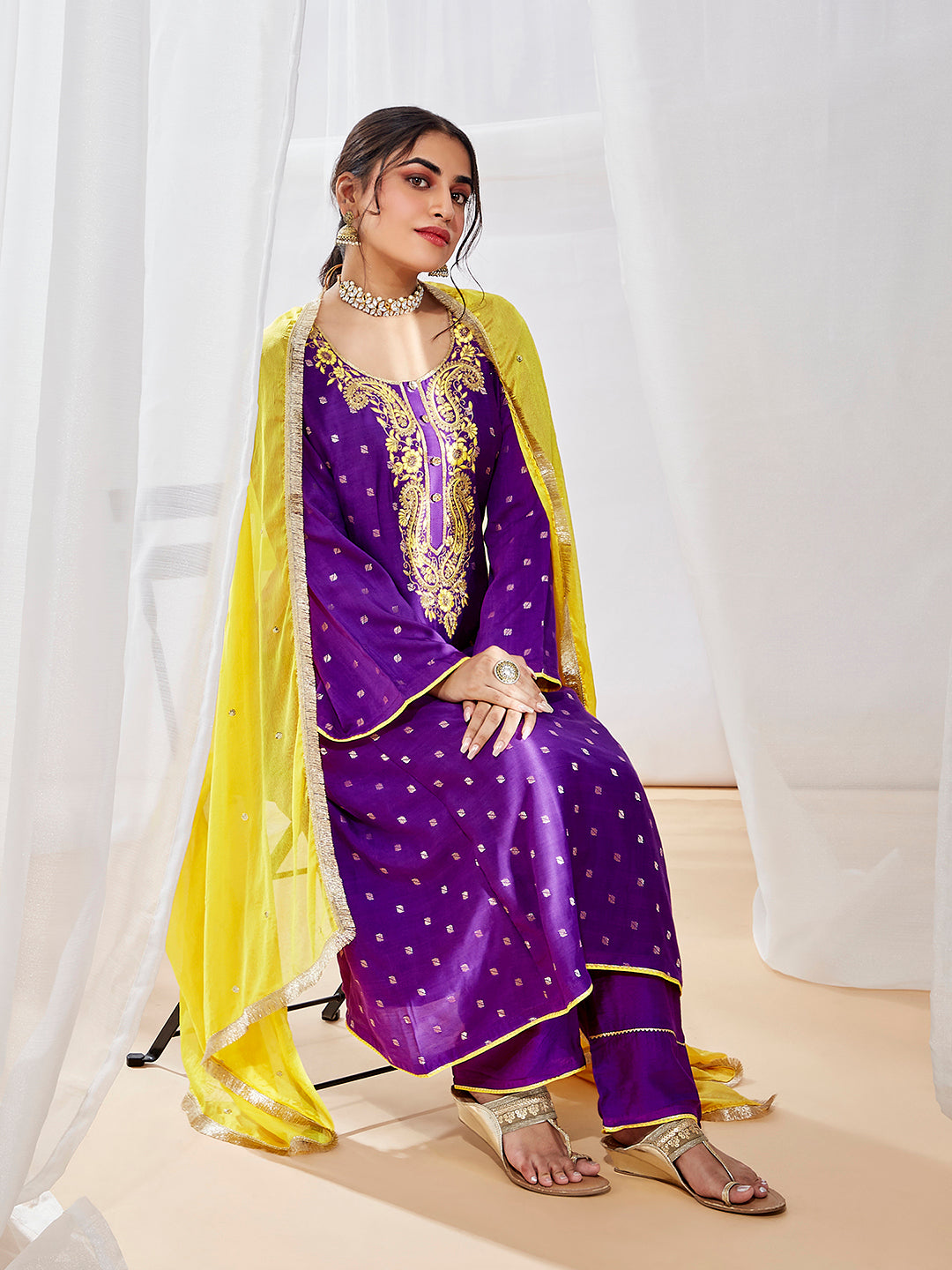A woman sitting, wearing a traditional purple kurta set with yellow dupatta and gold embellishments.